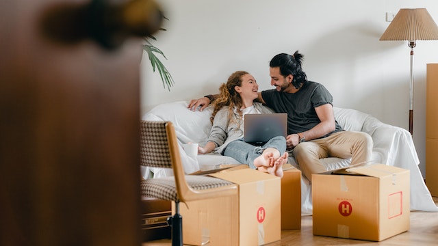 Smiling couple sitting on a couch looking at a laptop surrounded by moving boxes