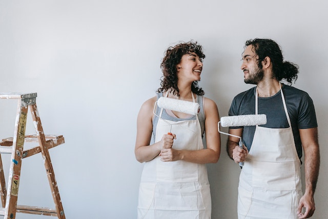 Two people wearing white aprons in a white room holding paint rollers and smiling at each other