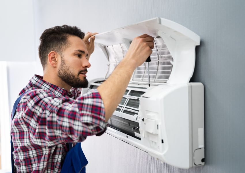 A property manager with a trimmed beard and  a flannel shirt inspects an air conditioner in their tenant's unit. 