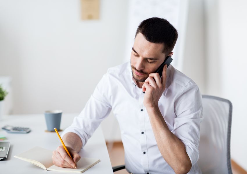 A property manager with dark short hair and a beard sits at a desk and writes in a notepad while talking to a tenant on the phone.