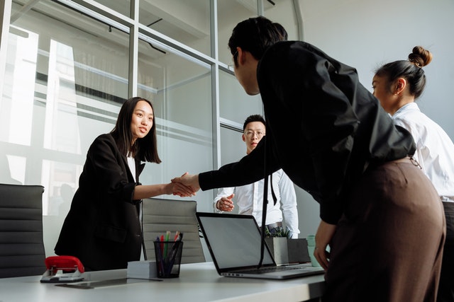 Four people meeting in an office, two of them are reaching across a table to shake hands