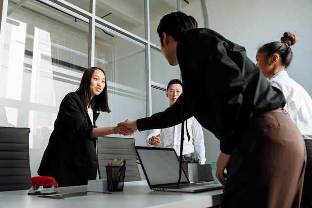 Four people meeting in an office, two of them are reaching across a table to shake hands