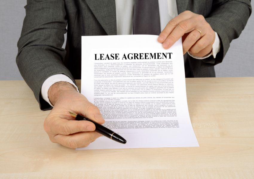 A landlord in a grey suit extends a black pen and lease agreement towards the camera, awaiting signature.