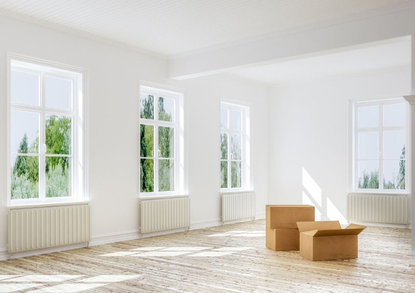 A white and light wood interior of a rental property with four bright windows is pictured on tenant move-out day, with two moving boxes on the bare floor.