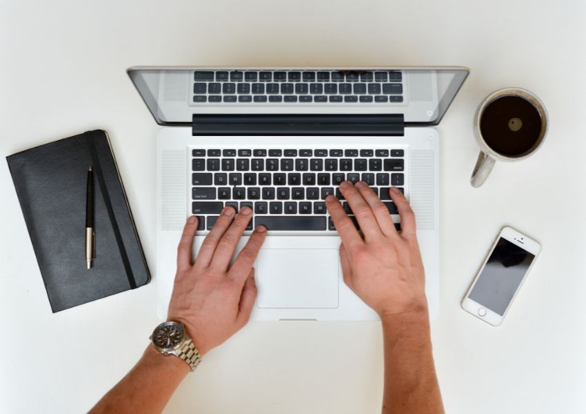 A landlord's hands are pictured drafting a move out letter on a silver and black laptop, with a black notebook, pen, coffee mug and phone on either side of the desk.