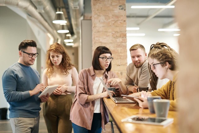 A group of six people leaning against a desk look at notebooks and computers and talking